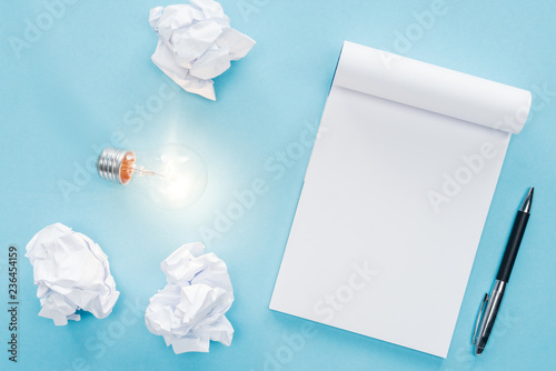 top view of blank notebook with crumbled paper balls and glowing light bulb on blue background, having new ideas concept