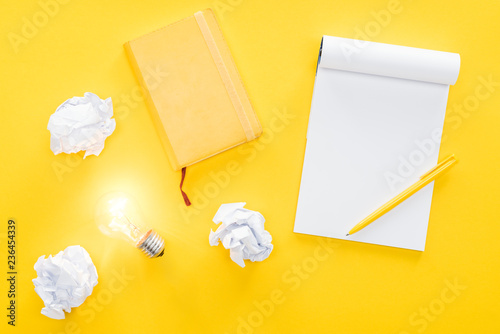 Obraz na plátně top view of blank notebook, crumbled paper balls and glowing light bulb on yello