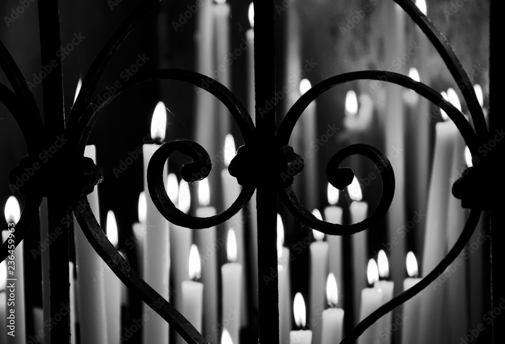 Candles light in dark temple seen through forging fence in heart shape. Religious background. Selective focus on the fence. Blurry lights. Black and white photo.