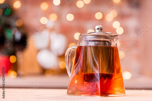 Conceptual transparent tea pot on a light wooden table against the background of the garland.