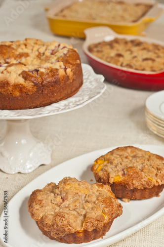 Top view, close distance of two, freshly baked homemade, peach streusel coffee cake on a round, white plate with other baked goods on display on a tan table cloth