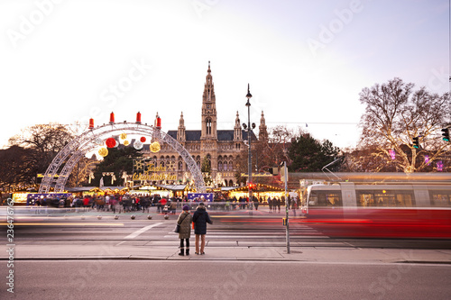 Vienna Rathausplatz and town hall at Christmas. People visiting the Christkindlmarkt, therefore waiting at traffic light to cross the road as blurred tram is passing by.