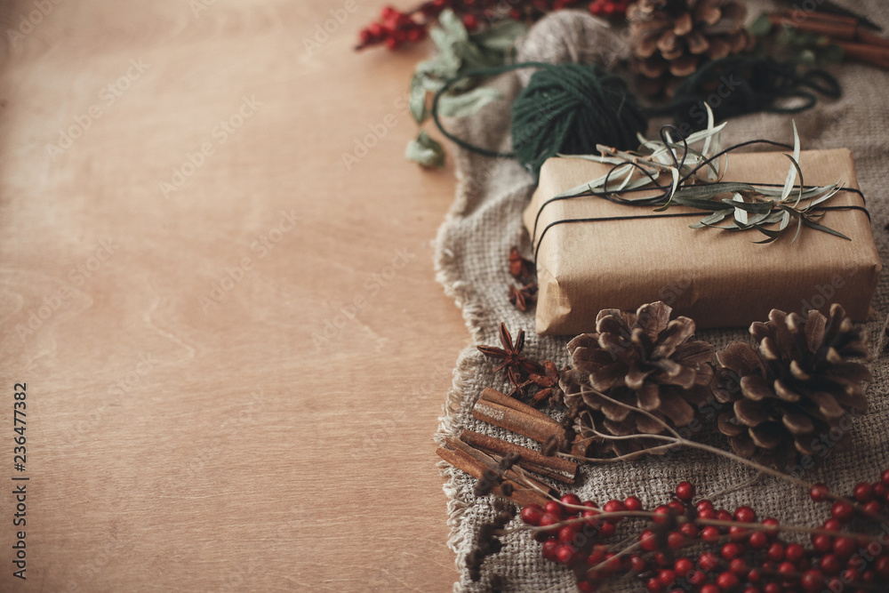 Fototapeta premium Stylish rustic christmas gift box with green branch and fir branches, berries, anise, pine cones, cinnamon on rustic wooden background. Simple eco present. Atmospheric mood. Space for text
