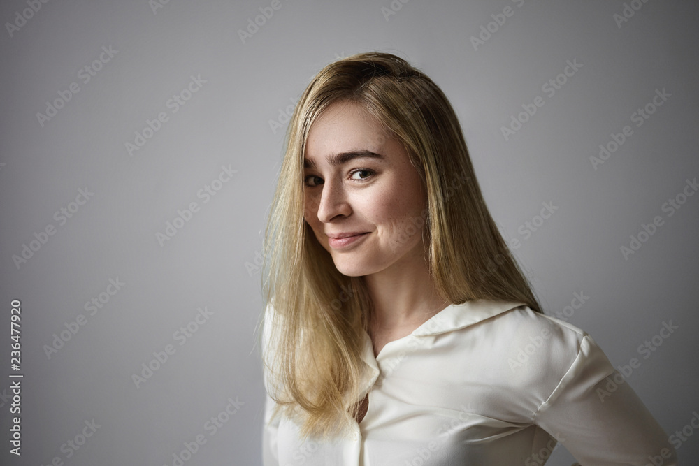 Cheerful mysterious young Europeanfemale with long blonde hair looking at camera with playful smile, having some secrets, being in good mood, posing isolated against blank gray wall background Cheerful mysterious young Europeanfemale with long blonde hair looking at camera with playful smile, having some secrets, being in good mood, posing isolated against blank gray wall background