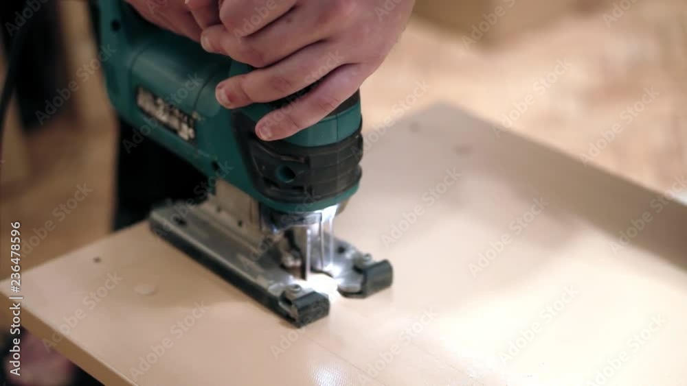 Closeup of hands with a manual electric jigsaw. Perform straight cut in a wooden surface. Closeup of hands with a manual electric jigsaw. Perform straight cut in a wooden surface.