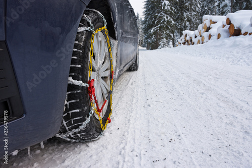 Snow chains mounted on front car tire in winter conditions with snow track and ice.