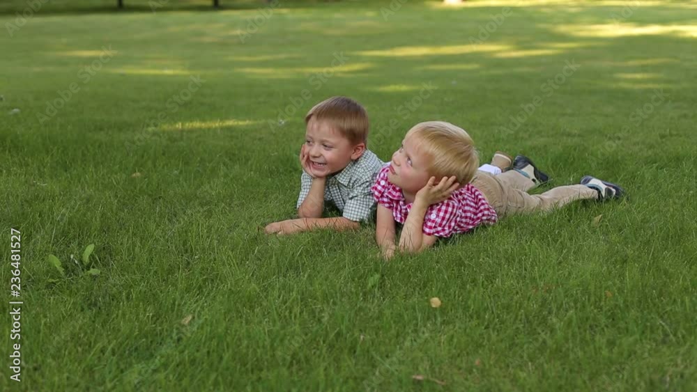 Two cute little boys in shirts lying on the green grass and smiling, portrait. Summer vacation. Young children relax lying on the grass in Sunny weather. Two cute little boys in shirts lying on the green grass and smiling, portrait. Summer vacation. Young children relax lying on the grass in Sunny weather.