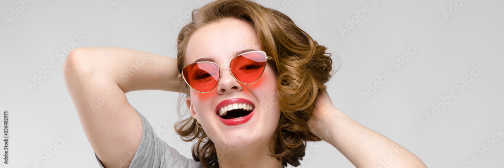 Charming young girl in a gray T-shirt on a gray background. Happy girl in red glasses. The girl put her hands behind her head Charming young girl in a gray T-shirt on a gray background. Happy girl in red glasses. The girl put her hands behind her head