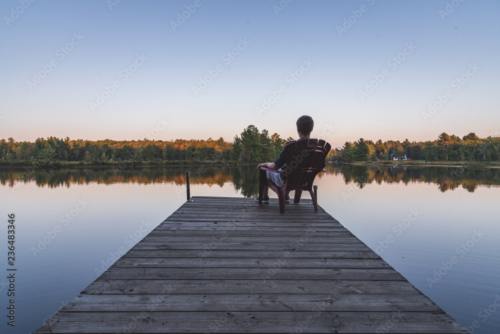 Young man relaxing on an Adirondack chair and looking at a calm river at sunset. Muskoka, Ontario, Canada.