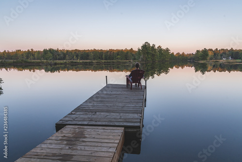 Young man relaxing on an Adirondack chair and looking at a calm river at sunset. Muskoka, Ontario, Canada. © Brian