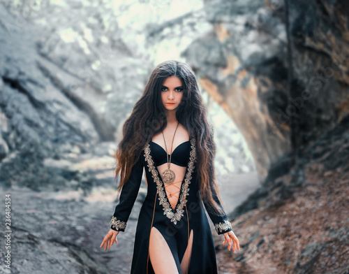 enchanted girl in a black dress with open tummy, bare legs and silver embroidery, with lush dark long hair and magical amulet around her neck. clairvoyant prepares a curse with a misty frozen forest