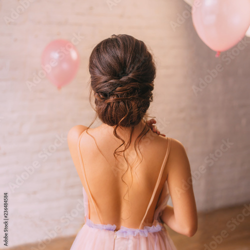 a young attractive lady, a peach dress with a purple color, shows a bare open back and a great neat dark hair hairstyle, standing in a bright spacious room with pink balls, no face in the photo