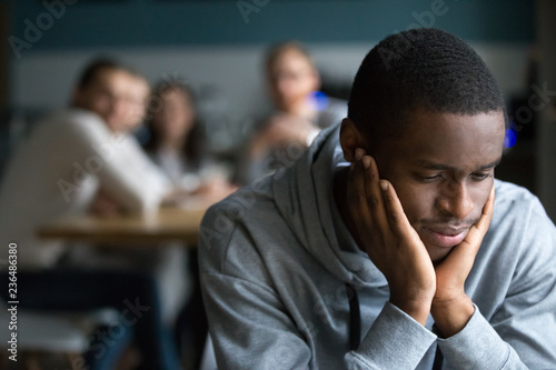 African American millennial guy feel lonely sitting alone in cafe, offended black student avoid talking to friends having misunderstanding, young man outcast suffer from racial discrimination