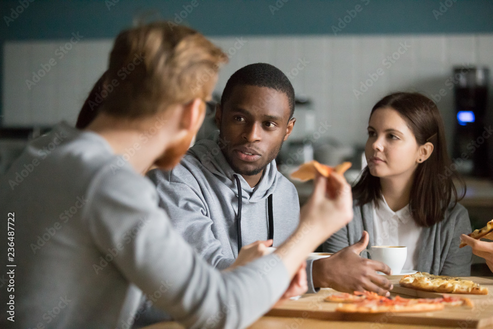 Multiethnic millennial colleagues talk meeting in pizzeria for lunch, diverse friends have conversation hanging out together in cafe, young people speak eating pizza, relaxing in Italian restaurant