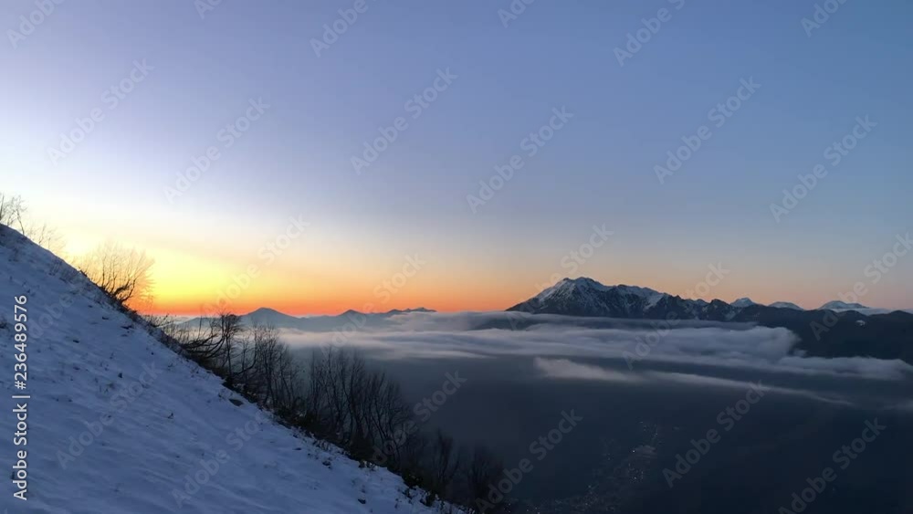 Golden evening sunset in sky over snowy mountain valley and lights modern city from drone. Aerial landscape evening sunset in cloudy sky and mountains valley. Drone flying over snowy mountains valley