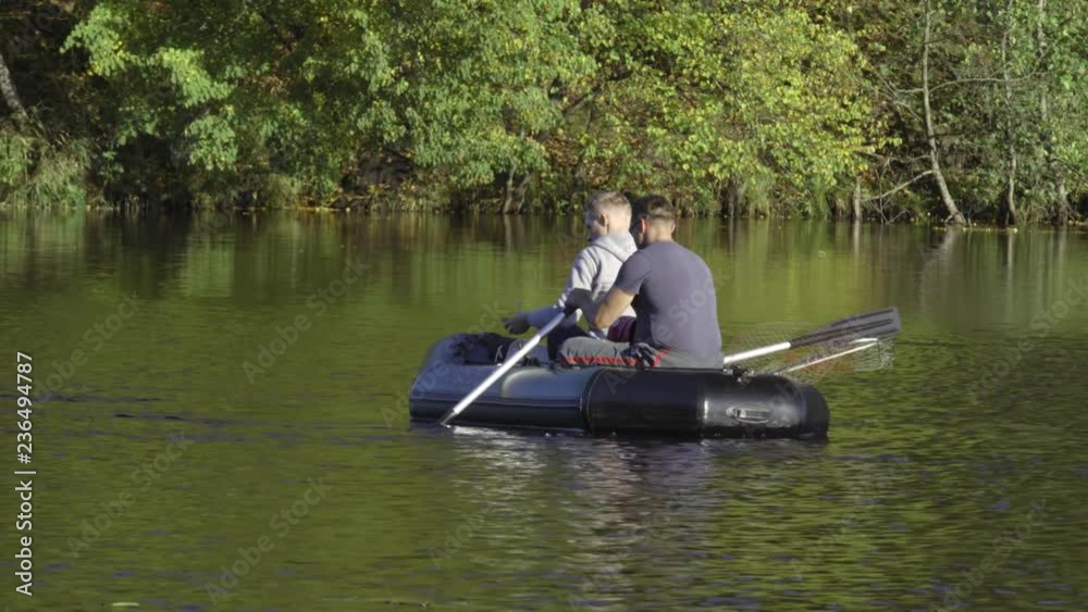 Two fisherman in rubber boat floats rowing with oars on river. Man is fishing on lake in sunny forest. Stracha river - beautiful place close to Belarusian nuclear power plant Two fisherman in rubber boat floats rowing with oars on river. Man is fishing on lake in sunny forest. Stracha river - beautiful place close to Belarusian nuclear power plant