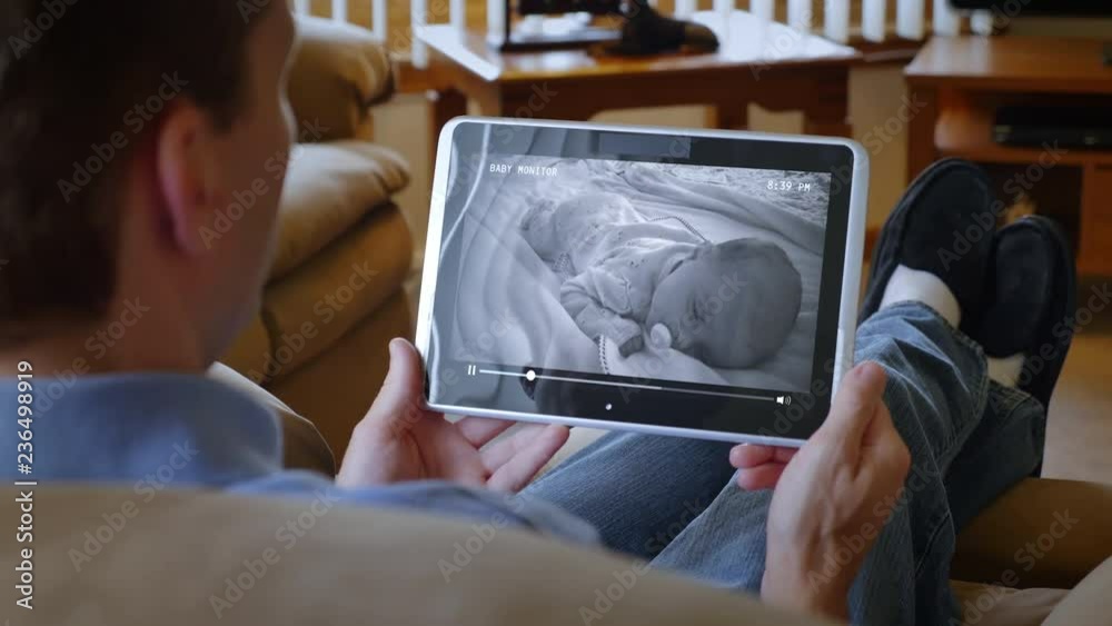 A father at home in his living room watches a baby monitor on his tablet PC. Black and white nightvision video of a sleeping infant on the screen.  	