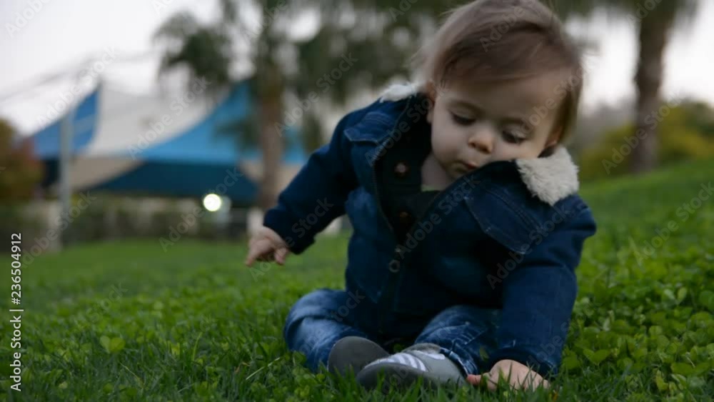 Portrait of cute stylish smiling toddler boy sitting on the grass in park outdoors. Positive smiling handsome blonde boy wearing blue jeans Portrait of cute stylish smiling toddler boy sitting on the grass in park outdoors. Positive smiling handsome blonde boy wearing blue jeans