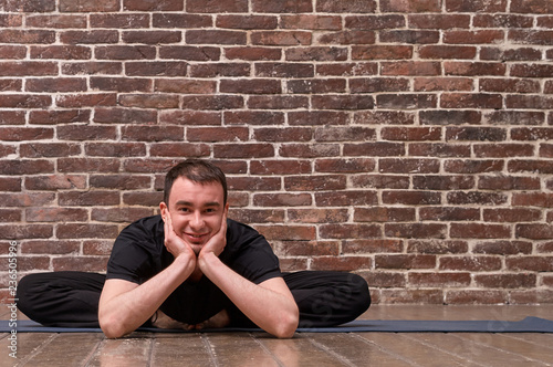 Sporty attractive young man working out, yoga, pilates, fitness training, asana Baddkha Konasana (Baddkhakonasana) over brick wall background. Studio shot. Full length photo