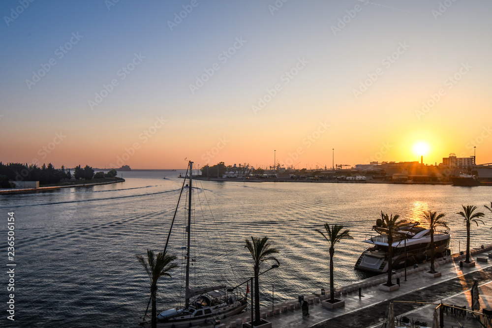 The sun sets over the harbor port as boats dock along the waterfront promenade in the coastal city of Brindisi, Italy, in the Puglia region