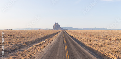 View of a road in a dry desert with a Shiprock mountain peak in the background during a vibrant sunny sunrise.Taken at Rattlesnake  New Mexico  United States.