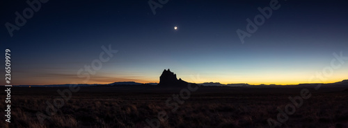 Dramatic panoramic landscape view of a dry desert with a mountain peak in the background during a clear night sky after sunset. Taken at Shiprock, New Mexico, United States.