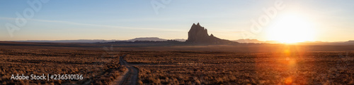 Striking panoramic landscape view of a dirt road in the dry desert with a mountain peak in the background during a vibrant sunset. Taken at Shiprock, New Mexico, United States.