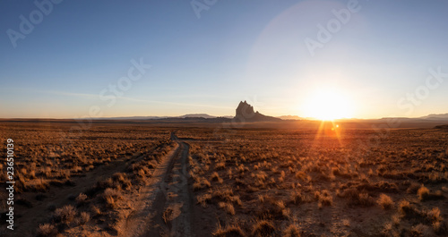Striking panoramic landscape view of a dirt road in the dry desert with a mountain peak in the background during a vibrant sunset. Taken at Shiprock, New Mexico, United States.
