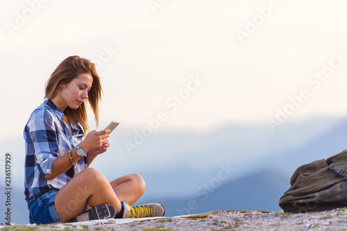 Fototapeta Naklejka Na Ścianę i Meble -  Young lady checking her phone and marveling the view from the top of the world at sunset. Success, winner, zen, tranquility.