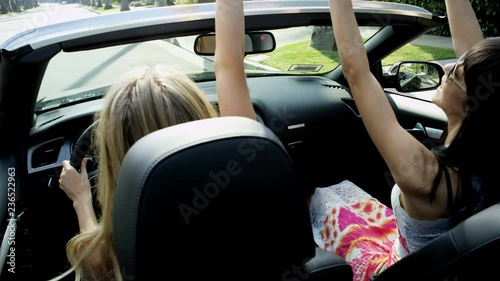Attractive European female friends singing, dancing and clapping driving luxury open top car along suburban street 