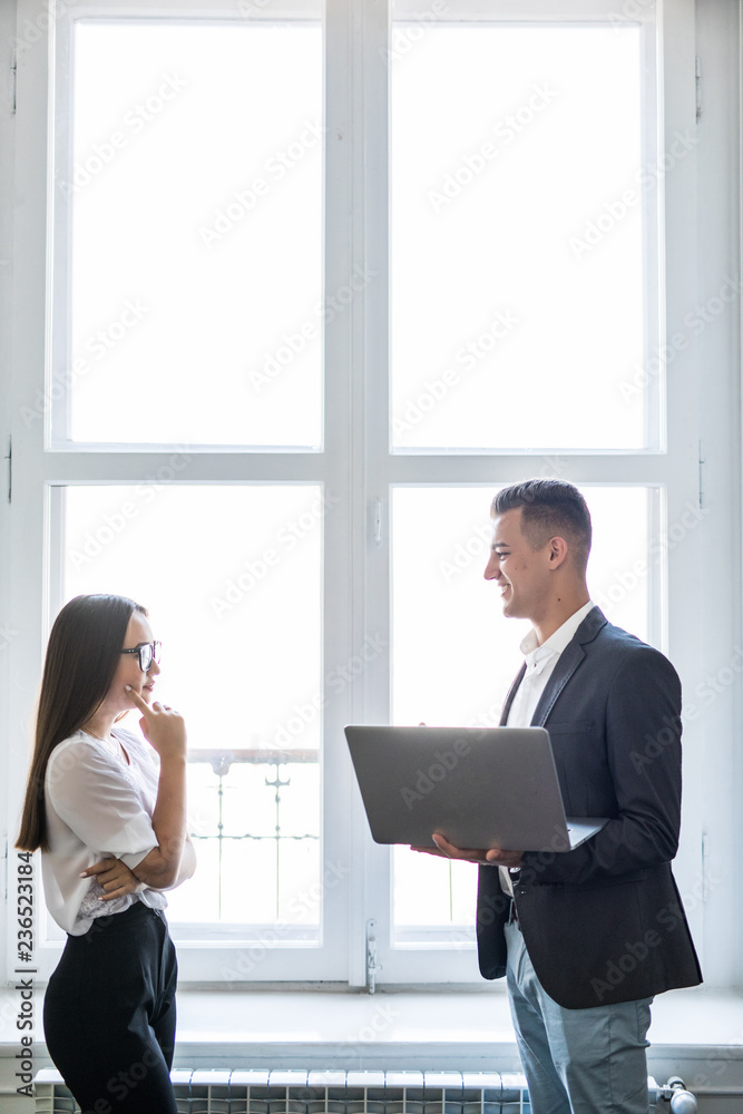 Businessman and asian businesswoman using a laptop together while standing in front of office building windows overlooking the city Businessman and asian businesswoman using a laptop together while standing in front of office building windows overlooking the city