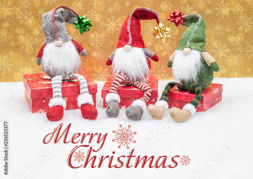 Weihnachtliche Wichtel mit Zipfelmützen sitzen auf Geschenken im Schnee mit goldenen Schneeflocken im Hintergrund, eine Grußkarte zur Weihnachtszeit.