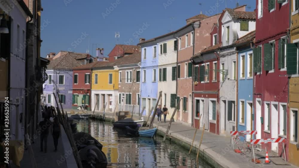 BURANO, ITALY - APRIL 21, 2018: Lovely colourful houses alongside the canal with moored boats and people walking. Scene of Burano island BURANO, ITALY - APRIL 21, 2018: Lovely colourful houses alongside the canal with moored boats and people walking. Scene of Burano island