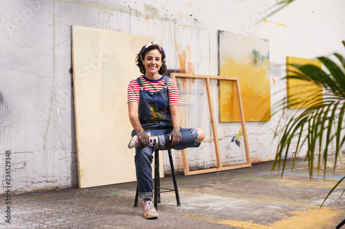 Female Artist Sitting With Paintings In Background At Studio
