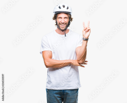 Handsome hispanic cyclist man wearing safety helmet over isolated background smiling with happy face winking at the camera doing victory sign. Number two.