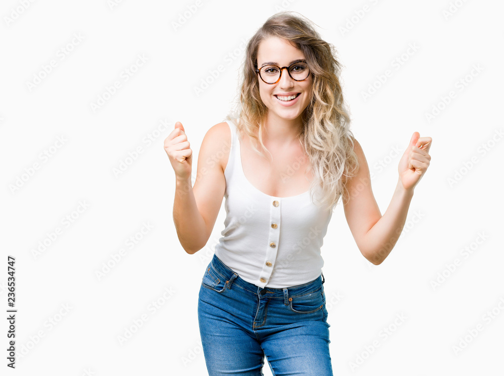 Beautiful young blonde woman wearing glasses over isolated background celebrating surprised and amazed for success with arms raised and open eyes. Winner concept. Beautiful young blonde woman wearing glasses over isolated background celebrating surprised and amazed for success with arms raised and open eyes. Winner concept.