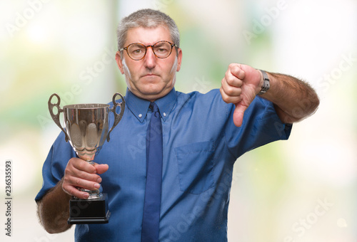 Handsome senior successful man holding trophy over isolated background with angry face, negative sign showing dislike with thumbs down, rejection concept