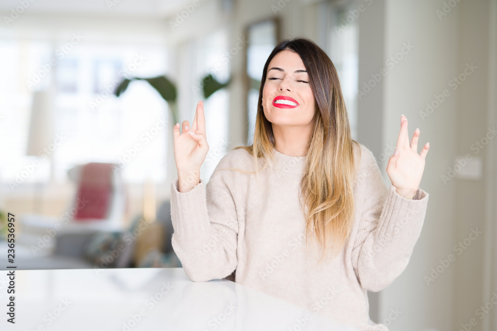 Young beautiful woman wearing winter sweater at home smiling crossing fingers with hope and eyes closed. Luck and superstitious concept. Young beautiful woman wearing winter sweater at home smiling crossing fingers with hope and eyes closed. Luck and superstitious concept.