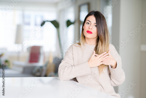Young beautiful woman wearing winter sweater at home smiling with hands on chest with closed eyes and grateful gesture on face. Health concept.