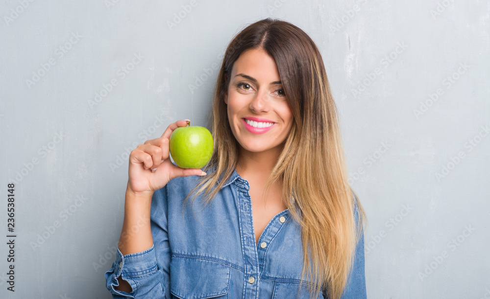 Young adult woman over grey grunge wall eating fresh green apple with a happy face standing and smiling with a confident smile showing teeth Young adult woman over grey grunge wall eating fresh green apple with a happy face standing and smiling with a confident smile showing teeth