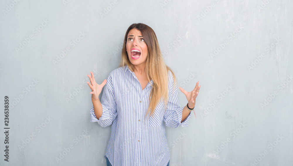 Young adult woman over grey grunge wall wearing fashion business outfit crazy and mad shouting and yelling with aggressive expression and arms raised. Frustration concept. Young adult woman over grey grunge wall wearing fashion business outfit crazy and mad shouting and yelling with aggressive expression and arms raised. Frustration concept.