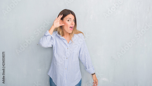 Young adult woman over grey grunge wall wearing fashion business outfit smiling with hand over ear listening an hearing to rumor or gossip. Deafness concept. © Krakenimages.com