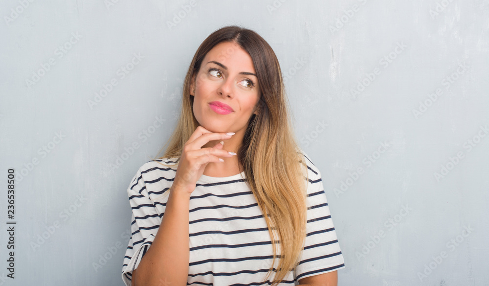 Young adult woman over grunge grey wall wearing navy t-shirt with hand on chin thinking about question, pensive expression. Smiling with thoughtful face. Doubt concept. Young adult woman over grunge grey wall wearing navy t-shirt with hand on chin thinking about question, pensive expression. Smiling with thoughtful face. Doubt concept.