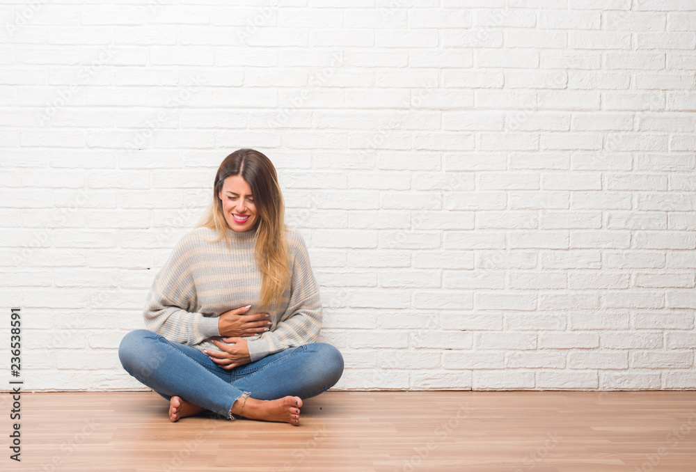 Young adult woman sitting on the floor over white brick wall at home with hand on stomach because indigestion, painful illness feeling unwell. Ache concept.