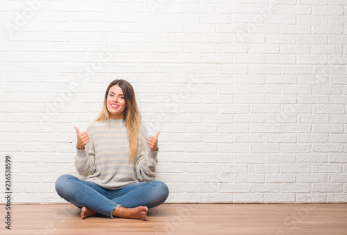Young adult woman sitting on the floor over white brick wall at home success sign doing positive gesture with hand, thumbs up smiling and happy. Looking at the camera with cheerful expression
