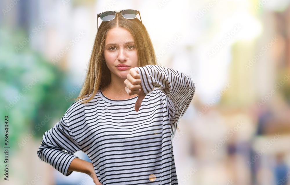 Young beautiful blonde woman wearing sunglasses over isolated background looking unhappy and angry showing rejection and negative with thumbs down gesture. Bad expression.