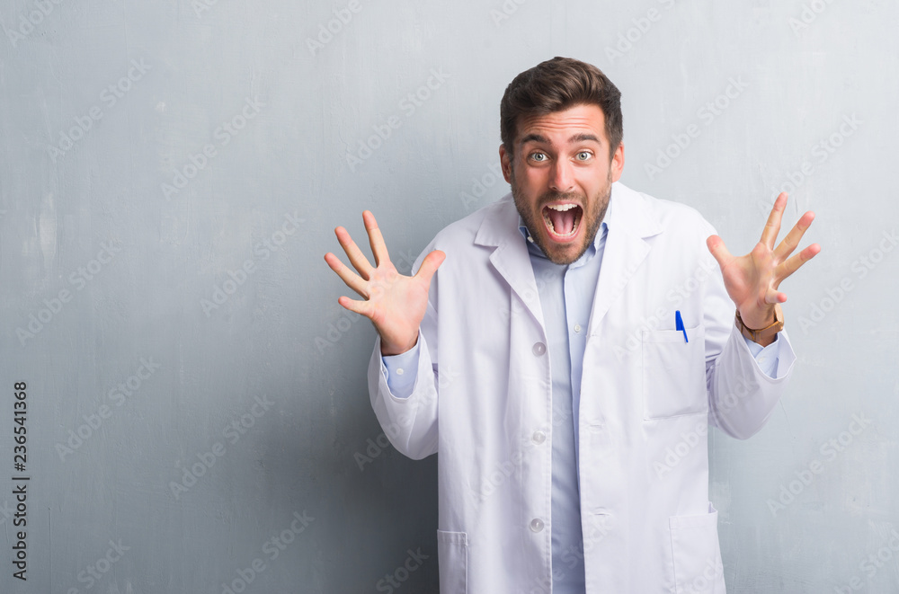 Handsome young professional man over grey grunge wall wearing white coat celebrating crazy and amazed for success with arms raised and open eyes screaming excited. Winner concept Handsome young professional man over grey grunge wall wearing white coat celebrating crazy and amazed for success with arms raised and open eyes screaming excited. Winner concept