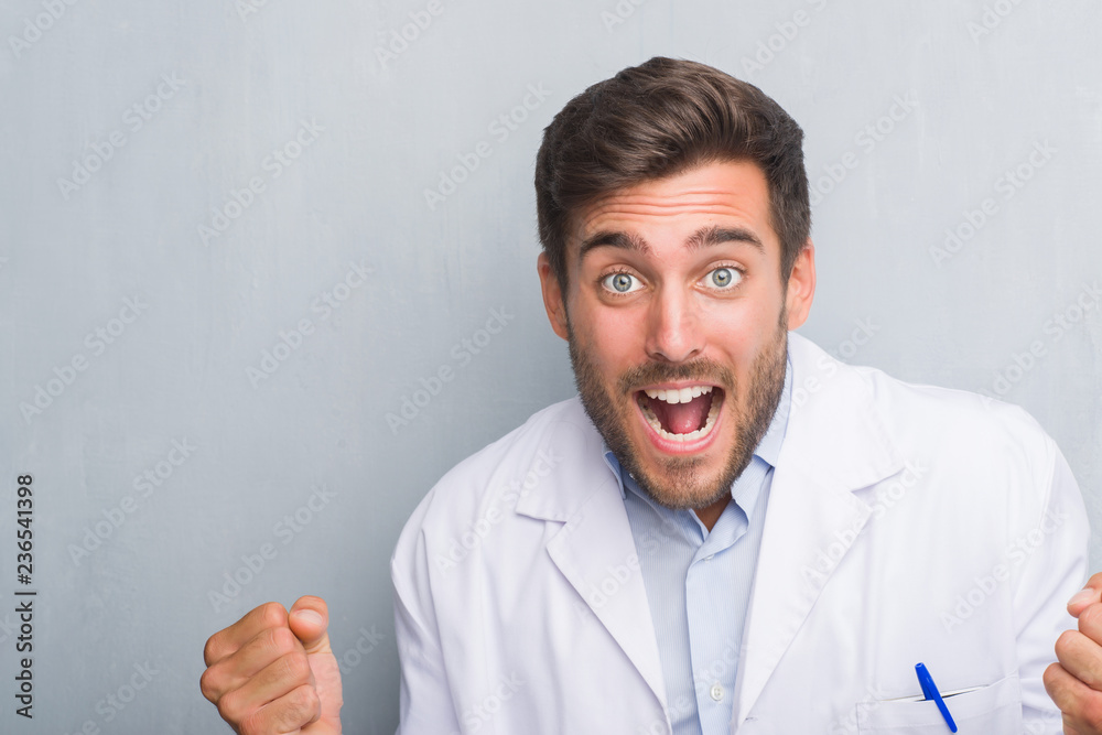 Handsome young professional man over grey grunge wall wearing white coat celebrating surprised and amazed for success with arms raised and open eyes. Winner concept. Handsome young professional man over grey grunge wall wearing white coat celebrating surprised and amazed for success with arms raised and open eyes. Winner concept.