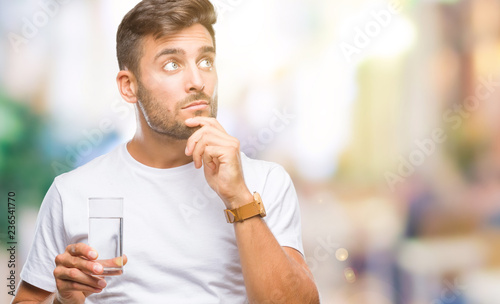 Young handsome man drinking glass of water over isolated background serious face thinking about question, very confused idea