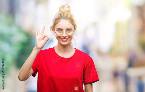 Young beautiful blonde woman wearing red t-shirt and glasses over isolated background smiling positive doing ok sign with hand and fingers. Successful expression.